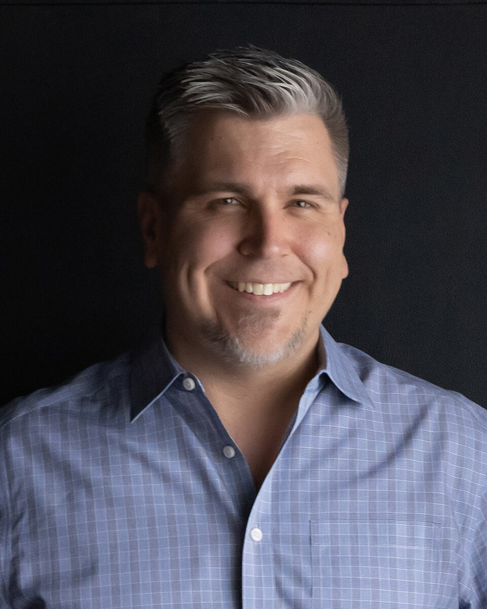 A man in a blue shirt smiling in front of a black background.