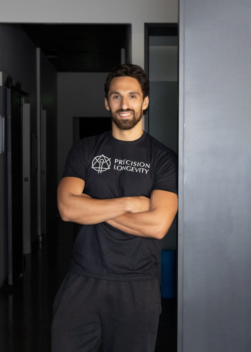 A man with a beard and mustache stands with arms crossed, wearing a black "Precision Longevity" t-shirt and dark pants, in a hallway with a gray wall and dim lighting.